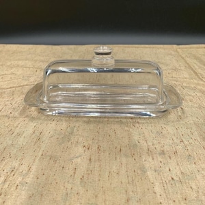 May include: A clear glass butter dish with a rectangular base and a domed lid. The lid has a small, round handle on top. The dish is sitting on a tan surface.