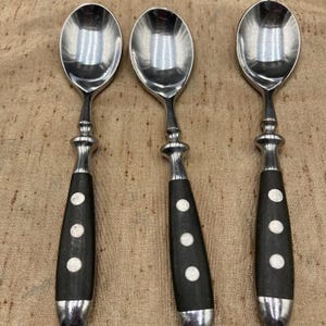 May include: Three silver spoons with black handles. Each spoon has a polished silver bowl, a decorative silver neck, and a black handle with three white dots. The spoons are arranged on a textured, tan surface.