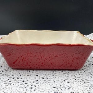 May include: A rectangular, red ceramic baking dish with a cream-colored interior. The exterior features a detailed embossed pattern. The dish has a scalloped rim and is suitable for baking casseroles and other dishes.
