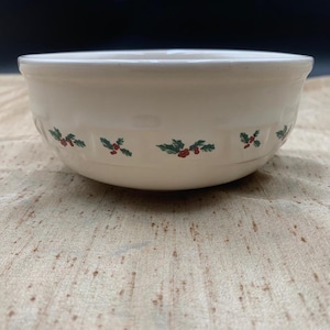 May include: A cream-colored ceramic bowl featuring a repeating pattern of green holly leaves and red berries around the exterior. The bowl has a smooth, rounded rim and a subtle embossed basket-weave texture along the upper portion of the side.