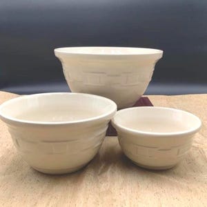 May include: A set of three cream-colored ceramic mixing bowls of varying sizes. Each bowl features a raised, embossed basket-weave pattern around the exterior. The bowls have a smooth, glossy finish and a wide, open circular rim design.