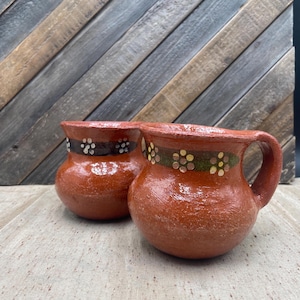 May include: Two terracotta-colored mugs with handles. Each mug has a decorative band of painted flowers and a dark brown band near the rim. The mugs are sitting on a light-colored surface with a wooden background.