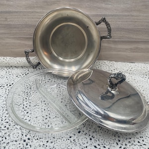 May include: Silver serving dish with a clear glass insert and lid. The dish has ornate handles and a decorative handle on the lid. The glass insert is divided into two sections. The dish is on a white lace tablecloth.