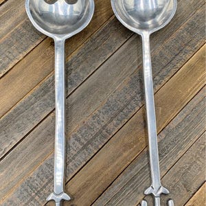May include: A pair of silver-toned metal salad serving utensils featuring long, straight handles that terminate in a decorative fleur-de-lis style base. One utensil is a solid round-bowled spoon, and the other is a slotted fork with three tines.