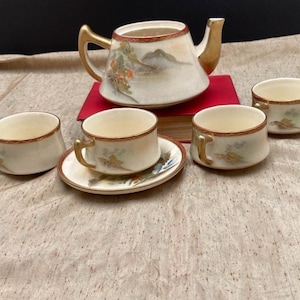 May include: A vintage Japanese tea set with a teapot and four cups. The teapot and cups are decorated with a floral and mountain landscape design in shades of gold, white, and brown.