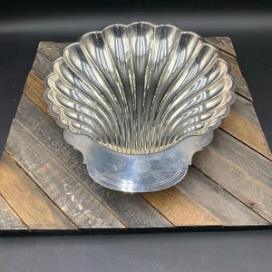 May include: A silver-colored, shell-shaped serving dish with a scalloped edge. The dish has a detailed, ribbed design and a flat, decorative base. The dish is placed on a wooden surface with various shades of gray and brown.