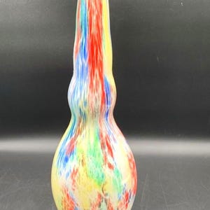 May include: A tall, colorful glass vase with a bulbous base and a slender neck. The vase features a vibrant mix of red, yellow, blue, and green hues, creating a speckled, abstract design. The vase is a decorative item.