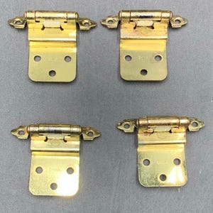May include: Four gold-colored cabinet hinges. Each hinge has a rectangular base with three holes and a curved top with two screw holes. The hinges are made of metal and have a shiny finish. The word "CHINA" is stamped on the base of each hinge.