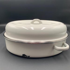 May include: A round, white enameled metal roasting pan with a matching lid. The pan features two side handles, a central handle on the lid, and a silver-toned rim. There is a small area of black enamel loss visible near the base of the pan.