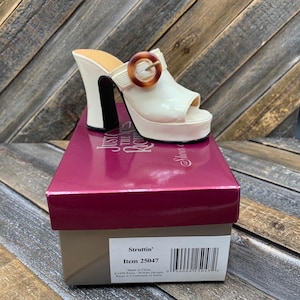 May include: A white platform sandal with a peep-toe design and a brown buckle. The heel is white with a black stripe. The shoe is displayed on a pink box with the text "Struttin'" and item number 25047.