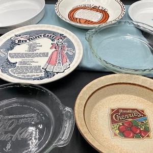 May include: A collection of various pie dishes and baking pans. Includes white ceramic, clear glass, and patterned ceramic dishes. One dish features a recipe for "Red Velvet Cake," and another displays a "Farm Fresh Cherries" design.