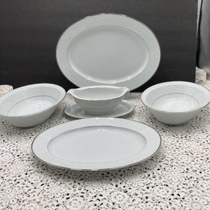 May include: A set of white porcelain dinnerware with a silver trim. The set includes a large oval platter, a smaller oval platter, two bowls, and a gravy boat with a matching saucer.