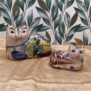 May include: Two ceramic cat figurines. The larger cat is beige with blue, yellow, and green floral designs. The smaller cat has similar colors and patterns. Both have black facial details and are on a tan surface.