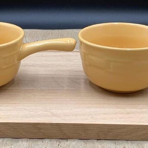 May include: Two matching light yellow ceramic bowls, each featuring a single side handle and a smooth, rounded interior. The bowls have a subtle, embossed brick-like pattern around the exterior walls and are presented together as a pair.