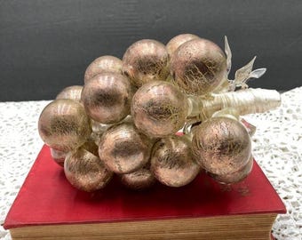 May include: A cluster of metallic gold-colored glass grapes with a textured surface, resting on a vintage red book. The grapes are attached to a clear stem with delicate leaves, creating a unique decorative piece.