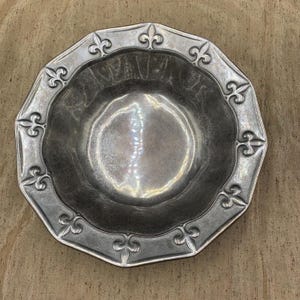 May include: A silver-colored, octagonal bowl with a scalloped rim. The rim is decorated with fleur-de-lis symbols. The bowl has a hammered texture and a reflective surface, suitable for serving or display.