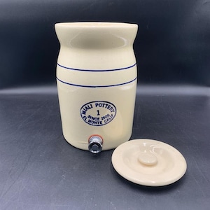 May include: Cream-colored ceramic water cooler with blue stripes and a spigot. The cooler has a lid and a circular emblem that reads "Miali Pottery Since 1906 El Monte Calif".