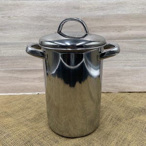 May include: A tall, cylindrical stainless steel stockpot with a polished, reflective finish. It features two curved side handles and a matching lid with a central arched handle on top. The metal surface shows clear reflections of the surrounding environment.