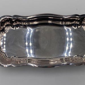 May include: A rectangular silver-toned serving tray with a polished, reflective surface and a scalloped edge. The tray features two ornate, curved handles on opposite ends, each decorated with intricate scrollwork and embossed detailing.