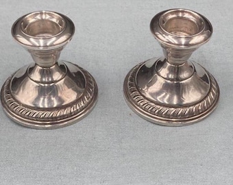 May include: A pair of polished silver-toned metal candle holders featuring a wide, circular base with a decorative rope-style border. Each holder has a smooth, tapered stem leading up to a circular cup designed to securely hold a single candle.