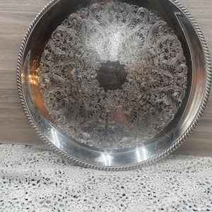 May include: A round, silver-colored serving tray with an ornate, floral pattern in the center. The tray has a rope-like border. The tray is likely made of metal and is suitable for serving food or displaying items.