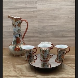 May include: A vintage porcelain tea set featuring a tall pitcher and four matching cups. The set has a white base with detailed illustrations of figures and landscapes, accented with red and gold trim. The cups are arranged on a mirrored tray.