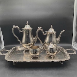 May include: A silver tea set on a rectangular tray. The set includes a teapot, coffee pot, creamer, and sugar bowl. The pieces have ornate handles and decorative detailing. The tray has handles on each side.