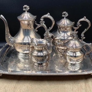 May include: A silver-toned tea service set consisting of a large teapot, a smaller lidded pot, a creamer, and a lidded sugar bowl. All pieces feature ornate handles and finials, arranged on a rectangular silver-toned serving tray with handles.