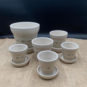 May include: A set of five off-white ceramic plant pots of varying sizes, each featuring a subtle horizontal band with a small dark green floral accent. Each pot includes a matching circular saucer base, arranged in a cluster on a textured surface.