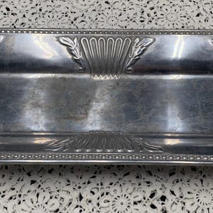 May include: A rectangular, silver-colored metal tray with an octagonal shape and ornate, raised floral and leaf designs. The tray has a shallow, recessed center and a beaded border. The overall design is vintage and decorative.