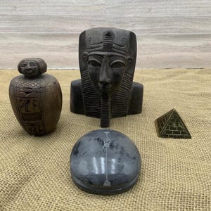 May include: A collection of Egyptian-themed decorative items. Includes a dark brown bust of a pharaoh, a small pyramid, a round container, and a small jar with a lid. The items are arranged on a tan burlap surface.