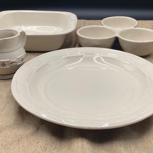 May include: A collection of cream-colored ceramic dinnerware including a large round dinner plate, a rectangular baking dish, a three-sectioned serving bowl, and a small pitcher with a subtle embossed grid pattern and small red floral accents.
