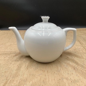 May include: A solid white ceramic teapot with a rounded body, a curved spout, a matching lid with a small knob handle, and a sturdy side handle. The teapot features a smooth, glossy finish and a clean, simple design for serving hot beverages.