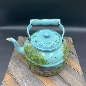 May include: A vintage, light blue tea kettle with a painted landscape scene. The kettle features a wooden handle and a lid adorned with floral accents. The landscape depicts a house, trees, and birds. The kettle is a charming piece of kitchen decor.