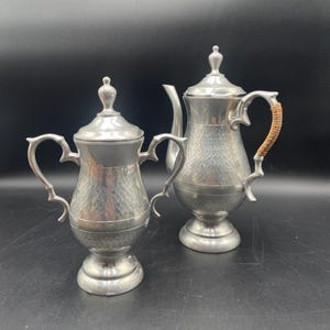 May include: Two vintage silver-toned metal coffee pots with lids. The pots have a hammered texture and ornate handles. One pot has a woven handle. The pots are on a black surface.