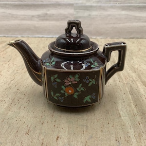 May include: A dark brown ceramic teapot with a lid and handle. The teapot features floral and leaf designs in shades of pink, green, and blue, with gold trim accents. The spout and handle are also dark brown.