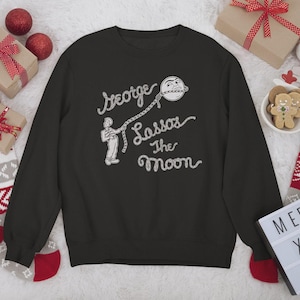 Pode incluir: Camisola preta com a frase "George Lassos The Moon" em uma fonte extravagante. O design apresenta uma figura de desenho animado laçando a lua. A imagem é cercada por decorações de Natal, incluindo presentes e enfeites.