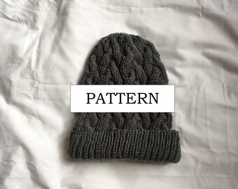 May include: A dark gray knitted beanie with a cable knit pattern. The hat has a folded brim and the word "PATTERN" is displayed on a white rectangle across the front. The hat is made of a soft, textured yarn.