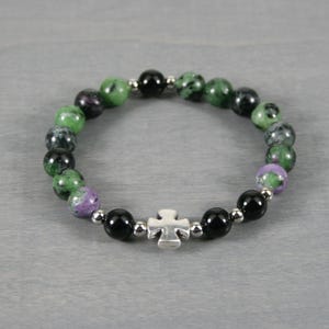 May include: A beaded bracelet featuring a silver cross charm. The bracelet is made of black, green, and purple gemstone beads, with small silver spacer beads. The bracelet is displayed on a gray surface.