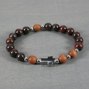 May include: A beaded bracelet featuring a silver cross charm. The bracelet is made with dark red and brown beads, interspersed with wooden beads and small silver-colored spacers. The beads are strung on an elastic band.