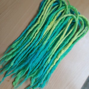 May include: A bundle of green and blue synthetic dreadlocks. The dreadlocks are made of wool and are ombre, transitioning from a light green to a dark blue.