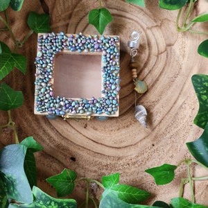 Op de afbeelding: Een kleine, vierkante houten doos met een helder venster, omlijst met iriserende, kiezelachtige stenen. Een decoratieve bedel met kralen, een zeeschelp en een zilveren spiraal hangt eraan. De doos staat tegen een houtnerf achtergrond, omringd door groene klimop.