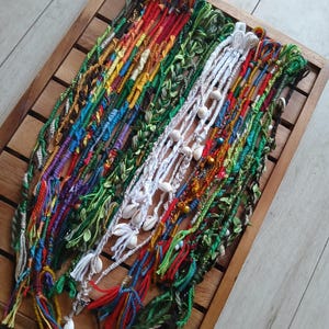 May include: A collection of colorful friendship bracelets with various beads and charms, including shells, tassels, and small leaves. The bracelets are displayed on a wooden tray.
