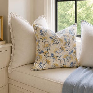 May include: A square throw pillow featuring a floral pattern with blue and yellow blossoms on a cream background, positioned next to a solid white pillow with a scalloped lace edge, both resting on a cushioned window seat with a blue throw blanket.