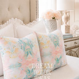May include: Two square throw pillows featuring a watercolor-style abstract pattern in soft shades of pink, teal, and yellow. The text DREAM DECOR IDEAS is printed in white on the front of the pillows, which are placed on a white quilted bedspread.