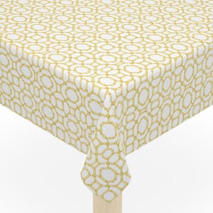 May include: A square table with light-colored wooden legs. The table is covered with a white tablecloth featuring a repeating gold geometric pattern. The tablecloth drapes over the edges of the table.
