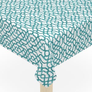 May include: A square white tablecloth with a teal, abstract, squiggly pattern covers a small wooden table. The table has four light-colored wooden legs. The tablecloth hangs over the edges of the table.