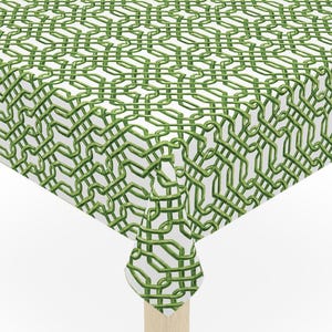 May include: A square white tablecloth with a green trellis pattern covers a small wooden table. The tablecloth has a repeating geometric design in shades of green. The table has four light-colored wooden legs.