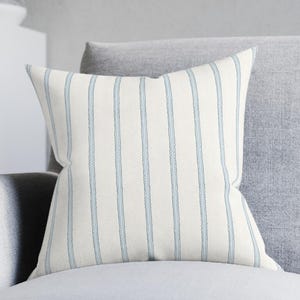 May include: A square throw pillow with a cream-colored background and vertical light blue stripes. The pillow is placed on a gray sofa, adding a touch of pattern and color to the neutral-toned living space. The pillow's design is simple and classic.