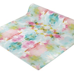 May include: A rectangular table runner featuring a soft watercolor-style print with blended hues of light blue, vibrant pink, and touches of yellow and green. The fabric is partially rolled at one end, displaying a repeating abstract floral pattern.